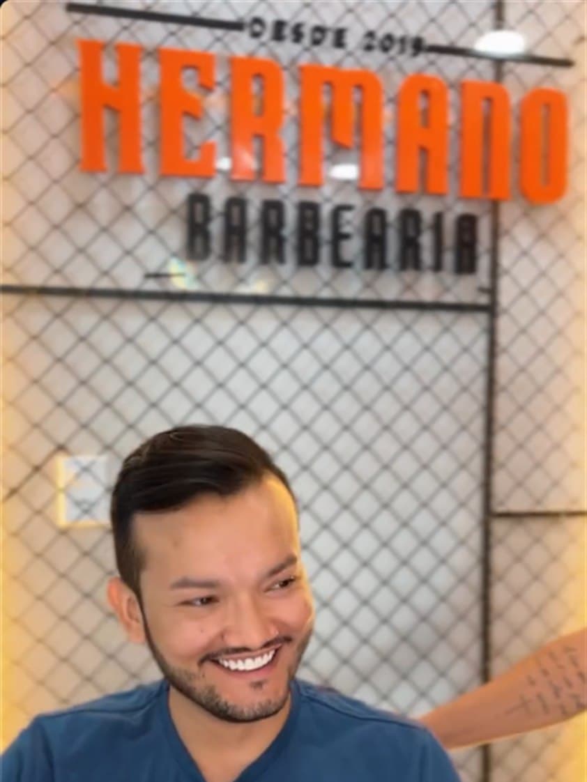 Cliente sorrindo diante da placa da Barbearia Hermano